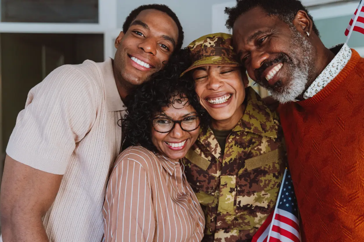 Four people are smiling and standing closely together. One person is in a military uniform, and another is holding small American flags.